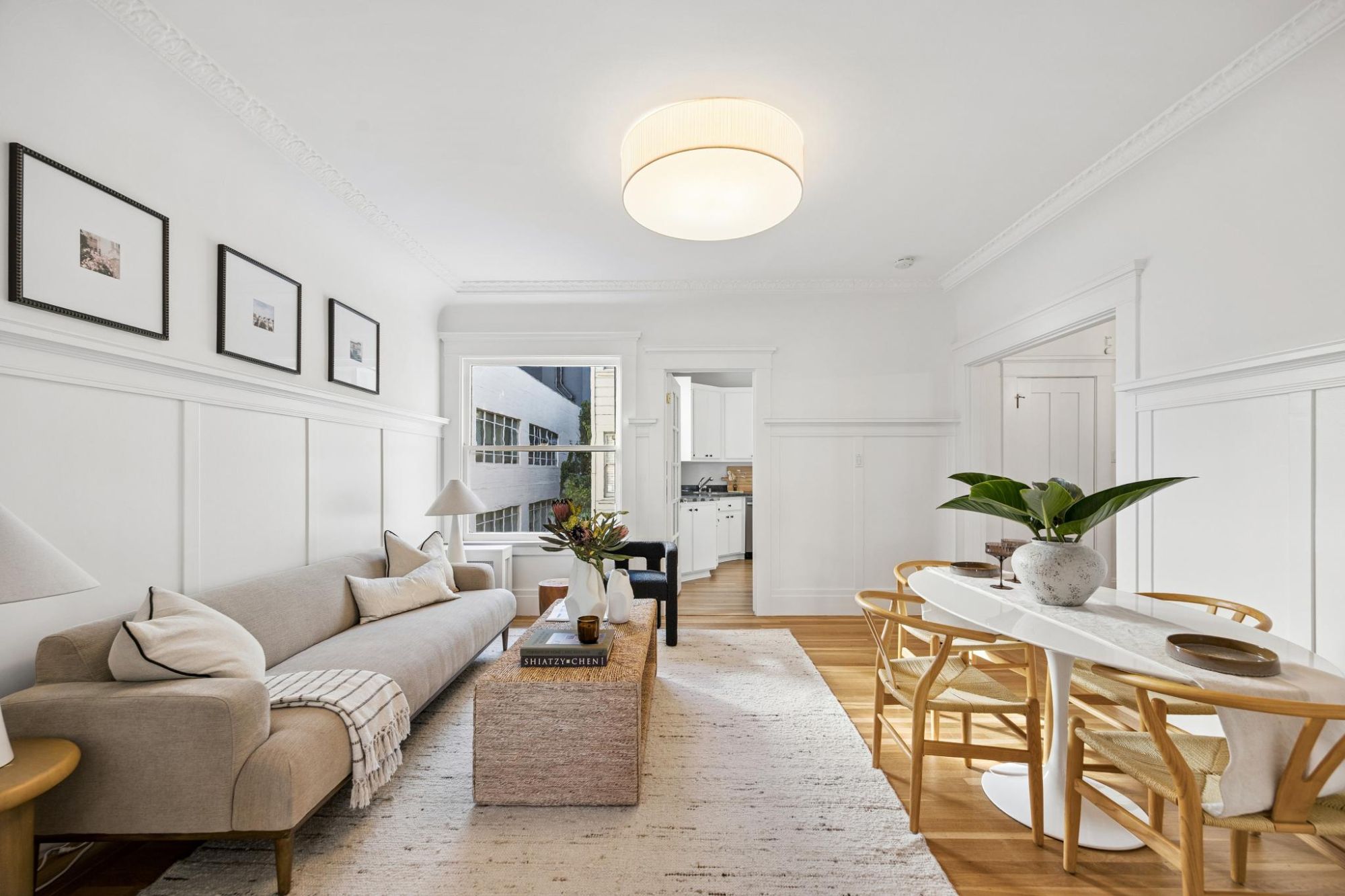 Living room with white wainscot paneling, ornate crown molding, a drum ceiling light, and a view through to the kitchen